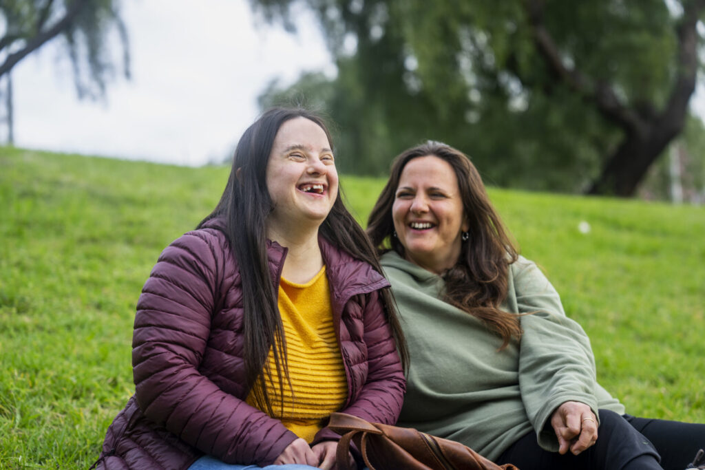 Woman with down syndrome laughing while sitting in grass with mother Woman with down syndrome laughing while sitting in grass with a friend