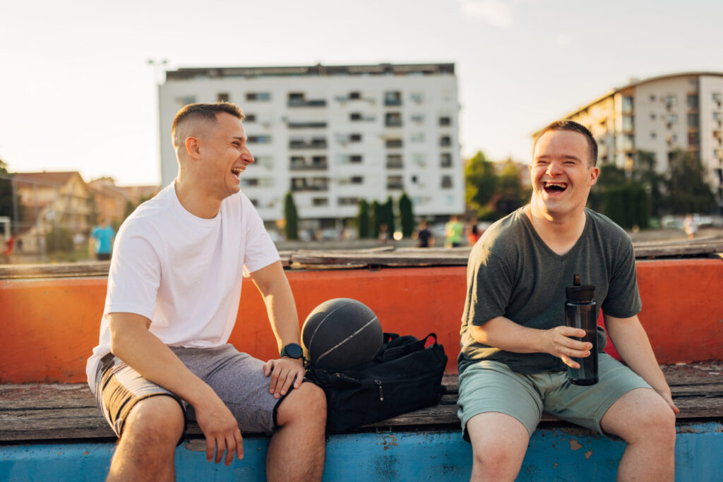 A smiling man with Down’s syndrome is sitting in the stands of the basketball court with his friend and having a great time A smiling man with Down's syndrome is sitting in the stands of the basketball court with his friend and having a great time